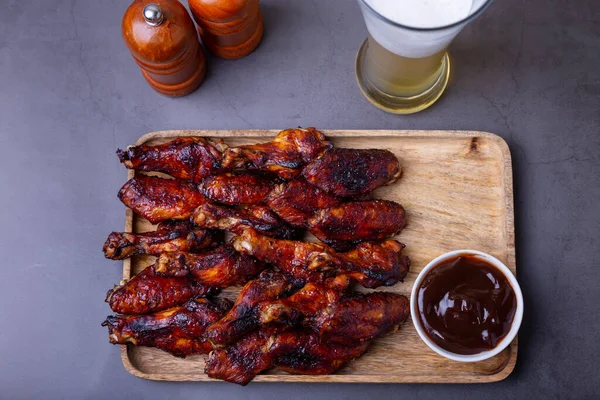 Ahşap bir tahtanın üzerinde kızarmış tavuk kanadı, barbekü sosu ve bir bardak bira. Siyah arkaplan, yakın plan.