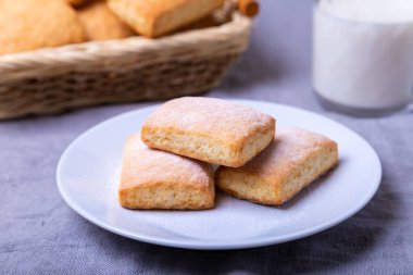 Beyaz tabakta pudra şekerli kurabiyeler. Arka planda bir sepet kurabiye ve bir fincan süt. Yakın plan..