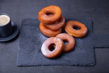 Pudra şekerli çörekler ve bir fincan kahve. Geleneksel donutlar, siyah arka planda yağda kızartılmış halka şeklinde. Abur cubur. Yakın plan..