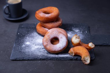 Pudra şekerli çörekler ve bir fincan kahve. Geleneksel donutlar, siyah arka planda yağda kızartılmış halka şeklinde. Abur cubur. Yakın plan..