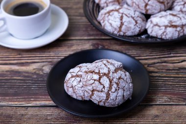 Çatlaklı çikolatalı kurabiyeler, üzerine şeker serpiştirilmiş. Ev yapımı pişirme, ahşap arka plan. Arka plan: bir fincan kahve ve bir tabak kurabiye. Seçici odak, yakın plan