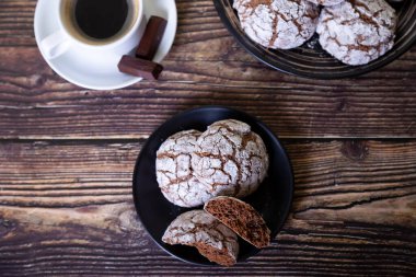 Çatlaklı çikolatalı kurabiyeler, üzerine şeker serpiştirilmiş. Ev yapımı pişirme, ahşap arka plan. Arka plan: bir fincan kahve, çikolata ve bir tabak kurabiye. Seçici odak, yakın plan.