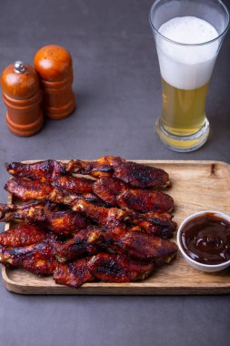 Ahşap bir tahtanın üzerinde kızarmış tavuk kanadı, barbekü sosu ve bir bardak bira. Siyah arkaplan, yakın plan.