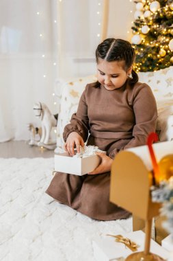Genç bir kız yumuşak bir halıya oturmuş, sıcak ve şenlikli bir oturma odasında hediye paketini dikkatlice açıyor. Tatil süsleri ve ışıklarla süslenmiş..