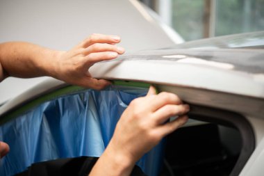 Bir oto tamirhanesinde araba penceresinin çerçevesine mavi maske bandı yapıştıran ve tamire ya da boyamaya hazırlanan ellerin yakın görüntüsü.