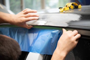 Kişi, ön uygulama için yüzeyi hazırlayan zımparalanmış bir alanın yanındaki bir araba kapısı paneline mavi koruyucu film uygular.