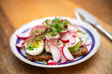 Dilimlenmiş yumurta turplu açık bir sandviçin yakın çekimi. Kalıplı bir tabakta taze otlar ve bulanık bıçaklı ahşap bir masa.