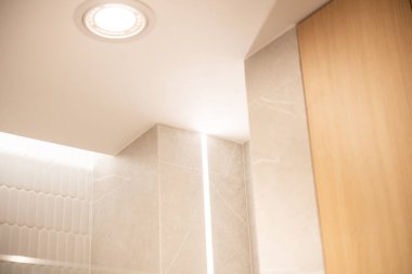 Modern bir banyo köşesinin yakın görüntüsü tenha tavan lambası, bej mermer fayanslar, dikey LED şerit lamba ve ahşap paneller..