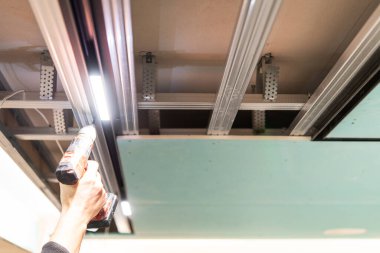 Modern bir iç mekanda askıya alınmış bir tavanın metal iskeletine LED ray ışıklandırması kurmak için kablosuz matkap kullanan el.