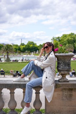 Güneş gözlüklü ve beyaz ceketli genç kadın güneşli bir günde Paris Fransa 'da Lüksemburg Bahçeleri' nde çeşmenin yanında dinleniyor.
