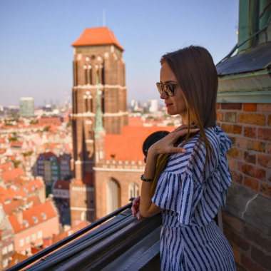 Güneş gözlüklü bir turist, elinde kamera ile çatı katında eski Gdansk kasabasını ve Polonya 'daki St. Marys kilise kulesini izliyor.