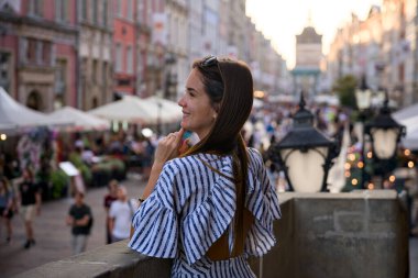 Meşgul eski kasaba caddesinin üstündeki yaya köprüsünde ayakta duran genç kadın altın saat boyunca Gdansk Polonya 'da kafe ve market tezgahları ile