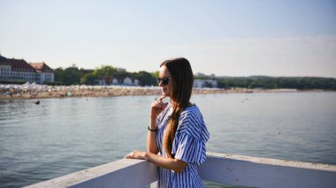 Polonya, Sopot 'ta deniz kenarındaki korkuluklara yaslanan güneş gözlüklü ve çizgili elbiseli şık bir kadın. Yaz tatili ve eğlence konsepti.