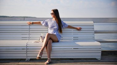 Yaz günü deniz kenarındaki beyaz bankta dinlenen genç bir kadın.