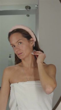 Kadın evinde sakin bir cilt bakımı sırasında aynanın önünde dururken boynunda gua sha aleti kullanıyor.