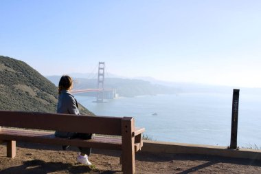 Genç bir kadın ahşap bir bankta oturuyor ve güneşli bir günde San Francisco, California 'daki Golden Gate Köprüsü' nün manzarasının tadını çıkarıyor.