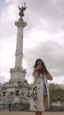 Kadın Bordeaux şehir meydanında bulutların altında heykel sütunuyla selfie çekiyor.