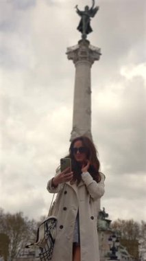 Genç kadın şehir meydanında yüksek anıtların yanında akıllı telefon özçekimi çekiyor.