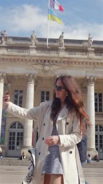 Bordeaux Fransız kadın güneşli bir günde 28 Mar 2025 'te şehir meydanında büyük neoklasik binaların önünde kolonlar ve bayraklarla selfie çekiyor.