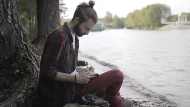 Человек делает заметки в блокноте, сидя на берегу — стоковое видео