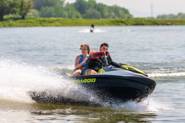 Krasnodar, Rusya - 25 Temmuz 2020: Arka koltukta mutlu bir kadınla erkek binici gün batımında kırsal kesimde nehir kıyısında su sıçrayan jet ski teknesi kullanıyor. Ekstrem sporlar ve yaz aktiviteleri konsepti