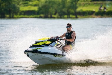 Krasnodar, Russia - July 25, 2020: BRP Sea-Doo jet ski watercraft driver in goggles drives water bike at high speed splashing at sunset by river bank. Active summer vacation concept clipart