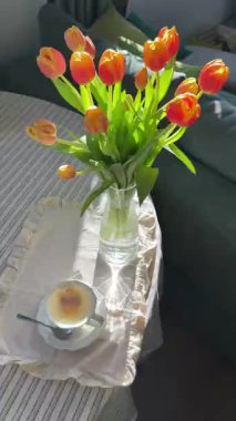 Parlak turuncu laleler açık bir vazoda, masanın üzerinde otururlar. Yanında kek olan bir fincan çay yakınlarda duruyor. Pencereden içeri güneş ışığı giriyor ve sıcak bir atmosfer yaratıyor..