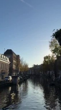 Amsterdam 'da insanlar kanal boyunca yürürken güneş ışığı suya yansıyor. Ağaçlar kıyılarda ziyaretçilerin dinlendiği ve şehrin manzarasını seyrettiği yerde..