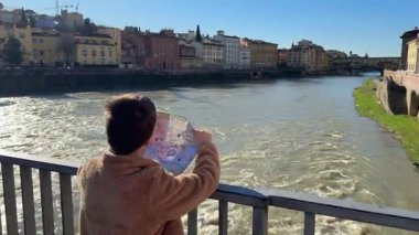 Bir çocuk Floransa 'da bir köprüde elinde bir haritayla nehre bakıyor. Manzara nehir kenarındaki binaları ve açık mavi gökyüzünü içeriyor..