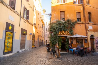 İnsanlar İtalya, Roma 'da bir kaldırım taşı sokağında bir barın dışında oturuyor. Ziyaretçiler konuşurken ve içkilerinin keyfini çıkarırken güneş batıyor. Yakınlardaki binaların parlak renkleri ve rahat bir havası var..