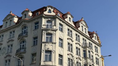 İnsanlar Prag 'da yüksek bir binanın yanında yürüyorlar. Yapıda ayrıntılı tasarım elemanları ve birden fazla balkon yer alıyor. Güneş ışığı sahnede parlıyor, mimariyle hava atıyor..