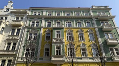 Prag caddesi boyunca yeşil, sarı ve beyaz dış görünüşlü binalar duruyor. Güneş parıldıyor, eşsiz mimari özellikleri vurguluyor..