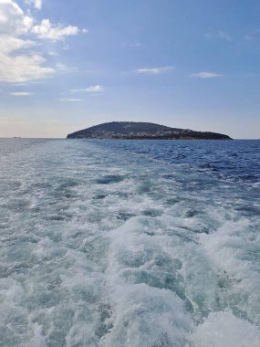 Hareket eden bir geminin bıraktığı köpüklü su izi ufuk çizgisindeki bulutlu gökyüzünün altındaki yeşil bir adaya doğru gidiyor.