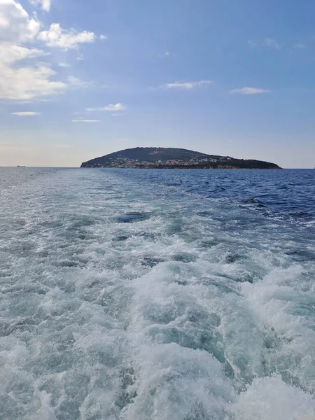 Hareket eden bir geminin bıraktığı köpüklü su izi ufuk çizgisindeki bulutlu gökyüzünün altındaki yeşil bir adaya doğru gidiyor.