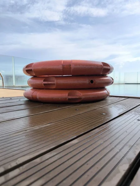 Üç parlak turuncu can yeleği nemli ahşap bir güvertede güzel bulutlu bir gökyüzünün altında berrak bir yüzme havuzunun yanında düzgünce istiflenmiş.