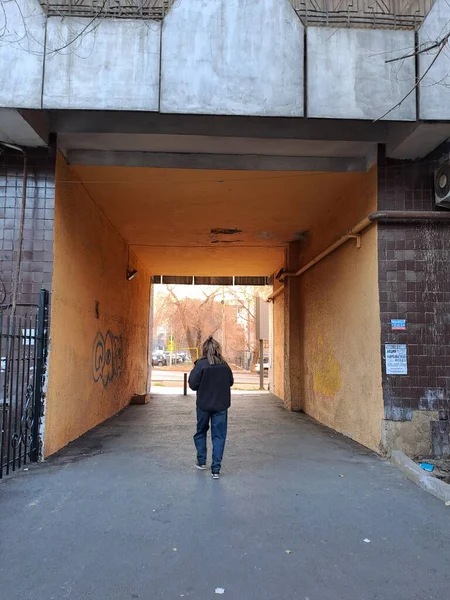 Koyu renk elbiseli bir insan, parlak sarı-turuncu duvarları olan bir geçitten geçerek ışığa ve dışarıda görünen çıplak ağaçlara doğru yürür.