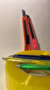 Keskin metal bıçaklı çoklu parlak plastik kullanım bıçakları bir rulo sarı paket bandına yerleştirilmiş. Ofis işleri, el işi, DIY projeleri ve tarafsız bir arkaplanda ambalaj için gerekli araçlar