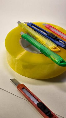 Keskin metal bıçaklı çoklu parlak plastik kullanım bıçakları bir rulo sarı paket bandına yerleştirilmiş. Ofis işleri, el işi, DIY projeleri ve tarafsız bir arkaplanda ambalaj için gerekli araçlar