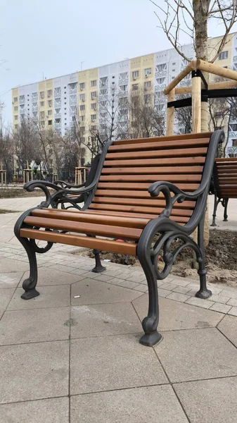 Kahverengi ahşap ve koyu metal çerçeveden yapılmış klasik park bankı kentsel yerleşim alanındaki bir kaldırımda duruyor.