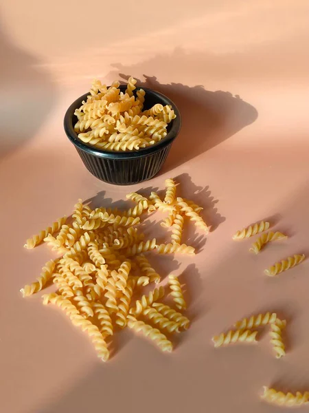 Şeftali rengi bir yüzeye saçılmış kuru fusilli makarna, siyah bir kasenin yanında.