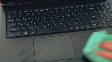 Toz ve kiri temizlemek için yumuşak yeşil bir bez kullanarak siyah bir dizüstü bilgisayarı ve dokunmatik pedi silen bir el.