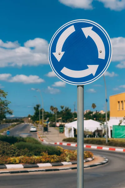 Close-up view of blue and white roundabout sign on the street with grass, trees, road and blue sky with clouds. Outdoor sign for cars. Traffic Laws. One circle road sign on a pillar.