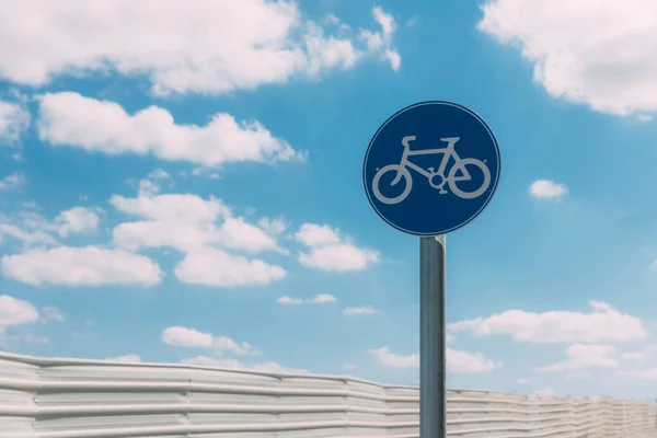 Close-up view of round blue and white bicycle lane sign against a fence and blue sky with clouds. Outdoor sign. Traffic Laws. One circle road sign on a pillar. Street signs. Safe bike rid