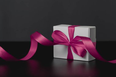 Beyaz hediye kutusunun yakın görüntüsü, koyu arkaplan ve yansımada lüks mor kurdeleli zarif bir kurdele. Mesaj için yeri olan hediye kutusu. Doğum günü, Noel, yıldönümü hediyeleri, hediye kartpostalları.