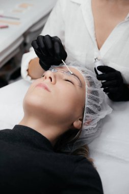 Kadının yüzüne yakın çekim, gözleri kapalı. Siyah eldivenli ustanın elleri, kalıcı makyaj prosedüründen önce kaş bölgesine pamuk çubuğuyla anestezi kremi sürerler. Güzellik Konsepti