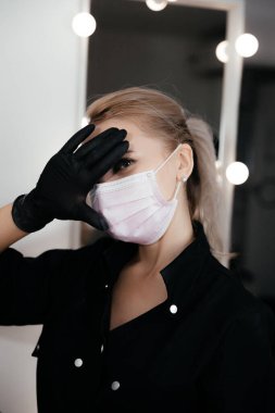Young permanent makeup master in light pink medical mask and black gloves with blurred mirror on background in salon. New fashion for beauty masters, cosmetologists in coronavirus epidemic period.
