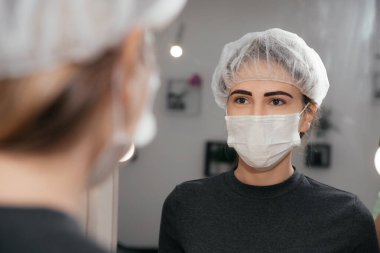 Kalıcı makyaj kaşlarından sonra tıbbi maske takan mutlu bir müşteri. Makyajlı bir kadın, makyajı bittikten sonra aynaya bakıyor. Güzellik salonu. Dövmenin sonucu. 