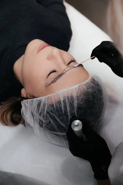 Close-up of the woman face with closed eyes. The hands of the master in black gloves applies anesthetic cream to the eyebrows before the permanent makeup procedure with a cotton stick. Beauty Concept