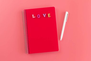 Kırmızı kapaklı, renkli harflerle kaplı, pembe arka planda aşk ve beyaz kalemle yazılmış bir defter. Sevgililer günü mesajı, aşk ve romantizm konsepti için hazırlan. Aşkıma mesaj yaz.