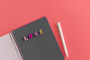 Renkli harflerle yazılmış gri defterli, açık spiral bir defter. Aşkla yazılmış ve pembe arka planda beyaz bir kalem. Sevgililer günü mesajı, aşk ve romantizm konsepti için hazırlan. İleti yaz